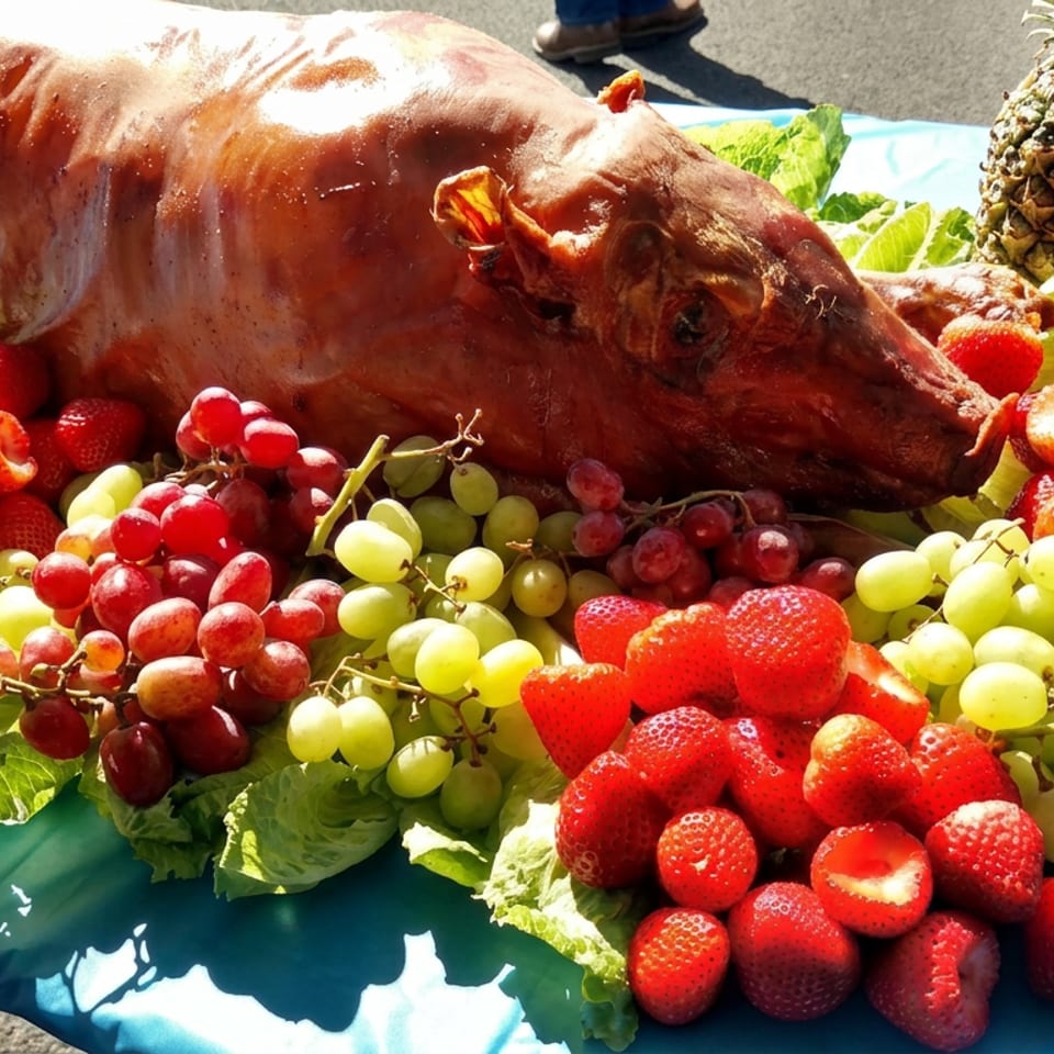 Roasted whole pig displayed on lettuce leaves, surrounded by strawberries and red and green grapes on an outdoor table.