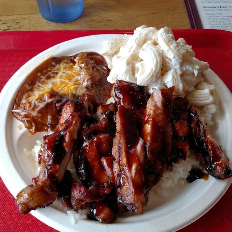 Plate of glazed grilled meat over rice with chili, shredded cheese, and creamy macaroni salad on a red tray.