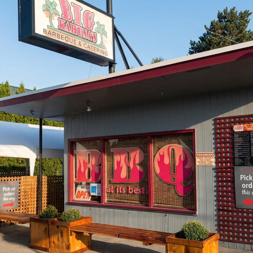 Big Kahuna’s Barbecue & Catering storefront with outdoor benches, pickup signs, and a white canopy under sunny skies.